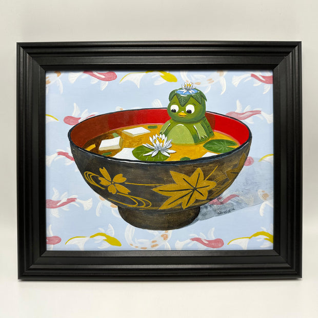 Painting of a small kappa, a green turtle like figure, sitting inside of a bowl of miso soup with cubes of tofu and small lily pads and a white flower. Background is a painted koi fish pattern. Piece is in black frame.