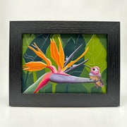 Illustration of a bird of paradise flower, with a small purple cartoon style hummingbird sitting at the end of its blooming. Behind is a background of solid green tropical leaves. Piece is in thick black wooden frame.