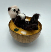 Needle felted sea otter laying in a wooden bowl filled with needle felted miso soup broth. Sea otter holds a tofu cube in its hands.