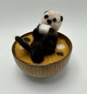 Needle felted sea otter laying in a wooden bowl filled with needle felted miso soup broth. Sea otter holds a tofu cube in its hands.