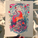 A hand holds a white art print featuring a vibrant, psychedelic illustration of a unicorn surrounded by mystical elements like a sword, stars, and red apples, set against a background of draped white fabric.
