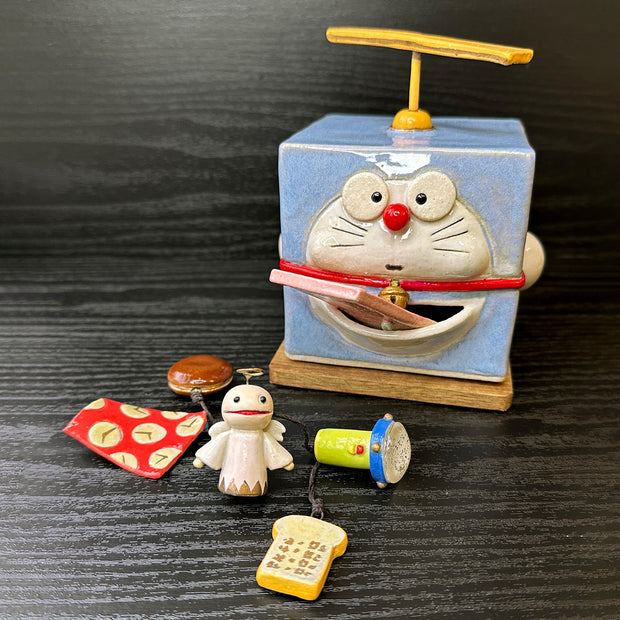 Ceramic cube designed to look like Doreamon, with a propeller coming out atop his head and a large square tongue coming out his mouth. In front of him is a collection of small trinkets, such as toast, an angel and flashlight.