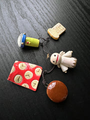 Chain of small trinkets made of ceramics, including: a flashlight, toast, an angel, a Dorayaki, and cloth with clocks on it.