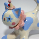 Ceramic figurine of a white cat with blue spots, wearing a pink ruffled collar, a small clown hat with clown makeup.