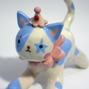 Ceramic figurine of a white cat with blue spots, wearing a pink ruffled collar, a small clown hat with clown makeup.