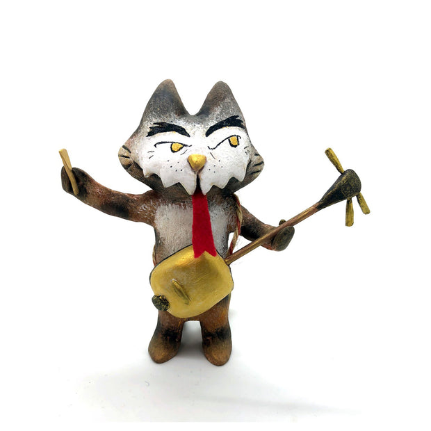 Painted sculpture of a cat with a white dripping face, gold eyes and nose. In one hand it holds the neck of a 3 stringed instrument slung around its body and the other holds a pick. A forked red tongue comes out of its mouth.