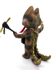 Painted sculpture of a cat with a white dripping face, gold eyes and nose. In one hand it holds the neck of a 3 stringed instrument slung around its body and the other holds a pick. It has 2 scaled tails.