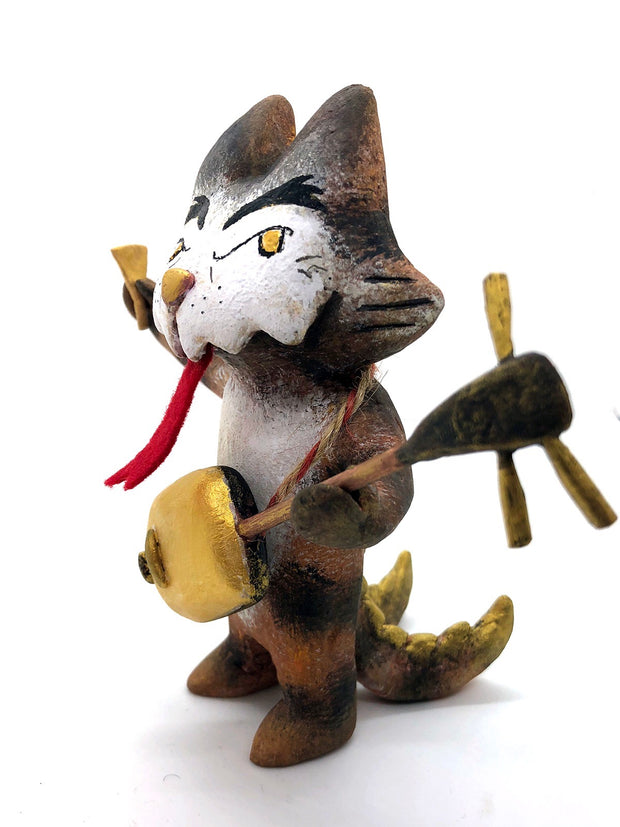 Painted sculpture of a cat with a white dripping face, gold eyes and nose. In one hand it holds the neck of a 3 stringed instrument slung around its body and the other holds a pick. It has 2 scaled tails.