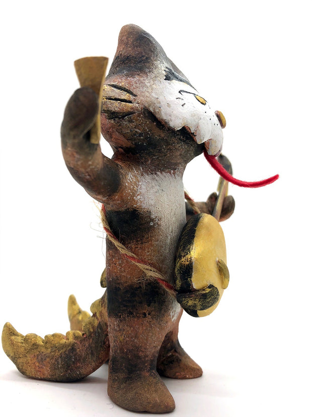 Painted sculpture of a cat with a white dripping face, gold eyes and nose. In one hand it holds the neck of a 3 stringed instrument slung around its body and the other holds a pick. It has 2 scaled tails.