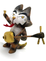 Painted sculpture of a cat with a white dripping face, gold eyes and nose. In one hand it holds the neck of a 3 stringed instrument slung around its body and the other holds a pick. It has 2 scaled tails.
