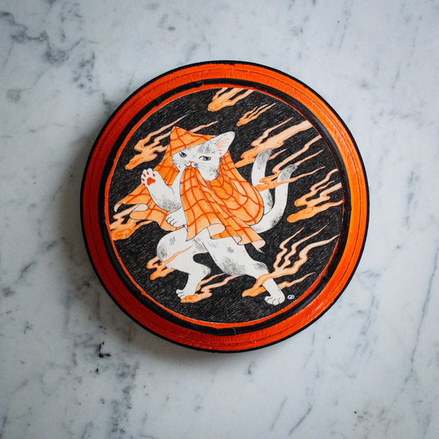 Black, white and orange illustration on a round orange and black panel. A white cat with 2 tails stands with a plaid blanket draped over its head and shoulders. It walks around orange wispy clouds.