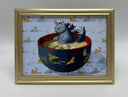 Framed artwork of a cat kaiju monster in a bowl of miso soup with a gold frame on a gray wall.