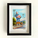 A framed artwork featuring a colorful illustration of a clown holding a 'Circus Liquor' sign on a street, mounted on a textured white wall.