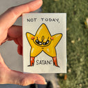 A hand holds an artwork of a yellow cartoon star character with the text 'NOT TODAY, SATAN!' in black lettering. The background shows an outdoor scene with pavement and grass.