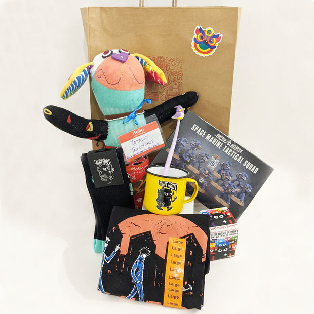 A cluster of items representing what comes in the lucky bag: multi-color plush dog with a name tag, black and teal giant robot socks, yellow camp mug, pen with a bird on the end, Space Marine Tactical Squad figure set, anime shirt in large, a big boss robot blind box. All of the items are in front of the Giant Robot retail bag with a Chinese new year sticker on the front