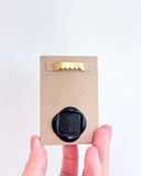 Hand holding a small beige panel with a black wax seal and gold sawtooth hanger on a light gray background