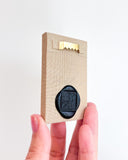 Hand holding a small beige panel with a black wax seal and gold sawtooth hanger on a light gray background