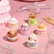 Small cake-shaped figurines on a pink surface with a birthday cake and decorations in the background.