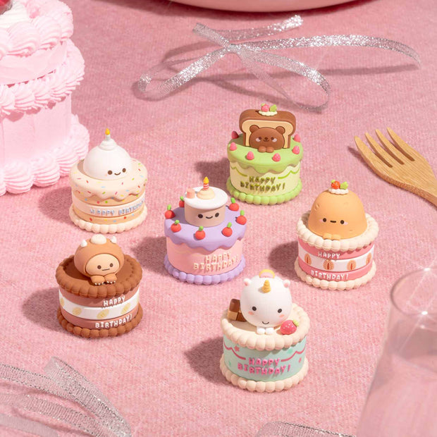 Small cake-shaped figurines on a pink surface with a birthday cake and decorations in the background.