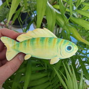 A hand holds a 6.75" long illustration of a green fish. It has striping down its body, large blue eyes, a wavy top fin, and a friendly, contented smile.