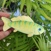 A hand holds a 5" long illustration of a green fish. It has striping down its body, large blue eyes, a wavy top fin, and a friendly, contented smile.
