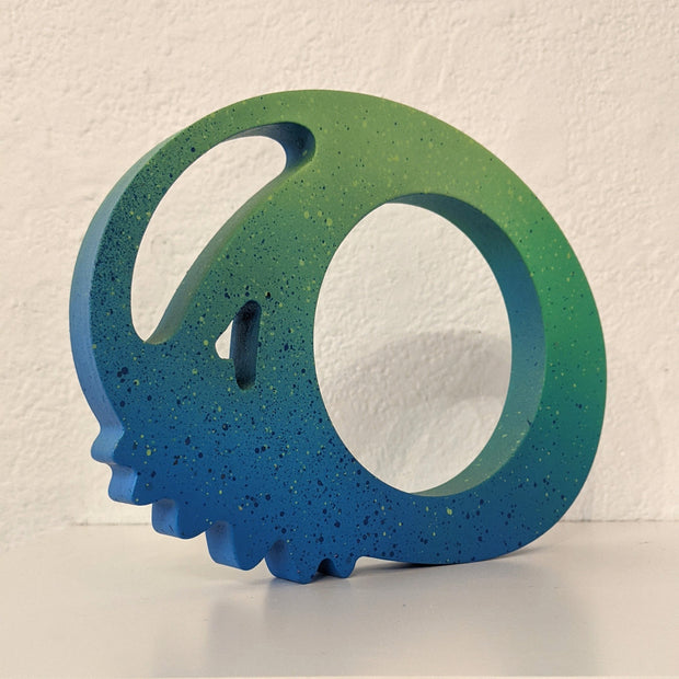 Green and blue wooden sculpture of a skull, with large eye sockets and rounded nubs for teeth.