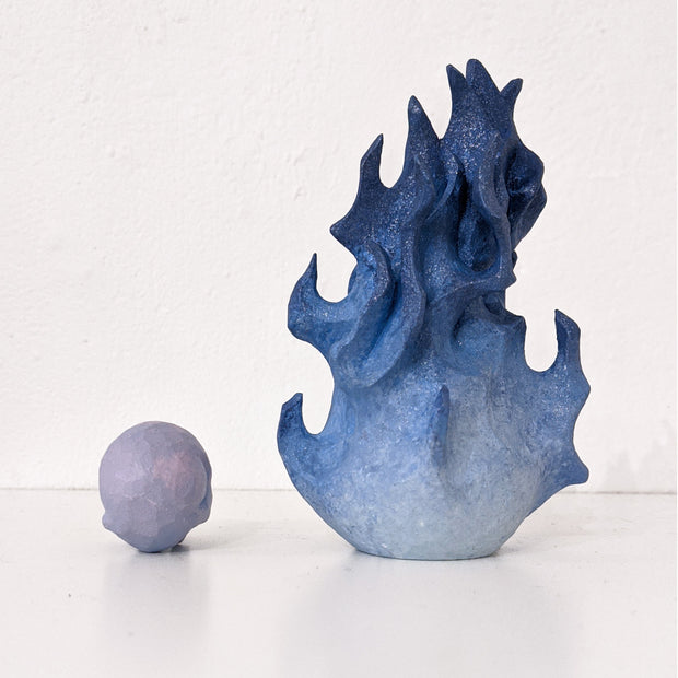 A small wooden sculpture of a purple skull and a taller sculpture of a blue flame with a closed eyed simplistic face. Viewed from behind.