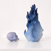 A small wooden sculpture of a purple skull and a taller sculpture of a blue flame with a closed eyed simplistic face. Viewed from the side.