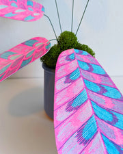 Sculpture of a plant made out of pink and blue risograph printed paper leaves, attached by wire to a pot. Leaves have a rounded striping pattern.