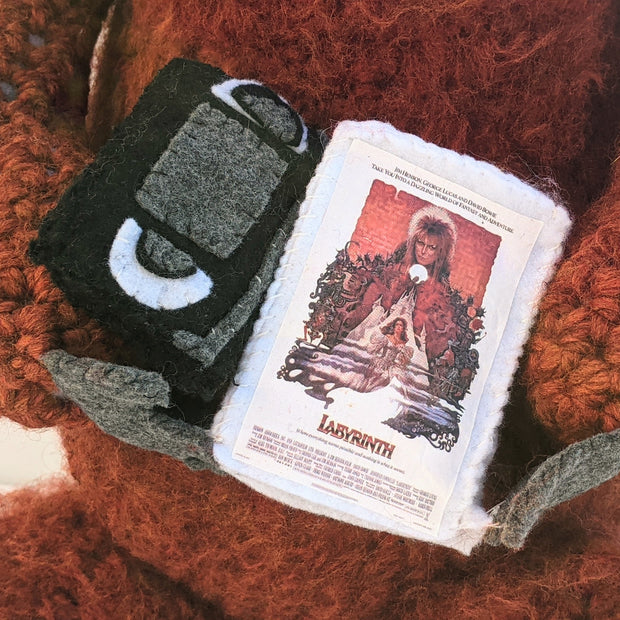 Large crocheted sculpture of Ludo from Labyrinth. He has messy hair and holds a copy of Labyrinth on VHS, which pops out of its case to reveal a crocheted disk.