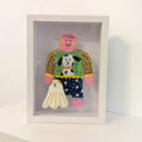 Paper cut out of a person wearing a shirt with patterning and a dog on it. Sitting at her feet is a long haired white dog.