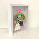 Paper cut out of a person wearing a shirt with patterning and a dog on it. Sitting at her feet is a long haired white dog.