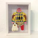 Paper cut out of a person wearing a shirt with patterning like a house with many windows. Sitting at their feet is a white dog with brown spots, made out of clay.