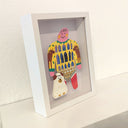 Paper cut out of a person wearing a shirt with patterning like a house with many windows. Sitting at their feet is a white dog with brown spots, made out of clay.