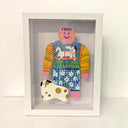 Paper cut out of a person wearing a shirt with patterning and skateboarding dog on it. Sitting at her feet is a white dog with brown and black spots.