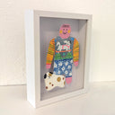 Paper cut out of a person wearing a shirt with patterning and skateboarding dog on it. Sitting at her feet is a white dog with brown and black spots.