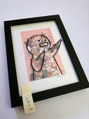 Framed artwork with a stylized character on a pink background with many, many cats on it., placed on a white surface.