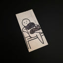 A rectangular drawing of a minimalist black ink illustration of a stick-figure-like figure holding a cat, photographed on a dark black background.