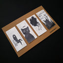 A set of four illustrated drawings on a brown kraft card against a black background. The postcards feature black and white hand-drawn illustrations, including a black cat, a small creature, and a dark landscape.