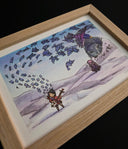 A framed artwork featuring a whimsical illustration of a small figure with a stringed instrument, floating paper coming off of it, a large gray statue, and a purple bird, displayed in a light wood frame against a black background.