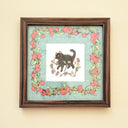 Framed artwork with a cat illustration and floral border on a beige wall.