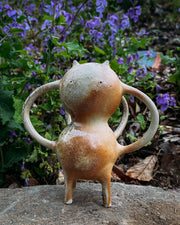 Brown ceramic sculpture of a character with a rounded body, small eyes and ears and mug handle like shapes coming out of its sides and back.