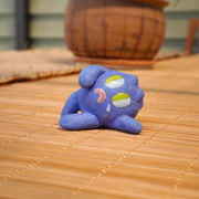 Small sculpture of a blueberry with a cute cartoon sad face - green eyes looking to the side and a frown with a single tear falling down. It lays on its side.
