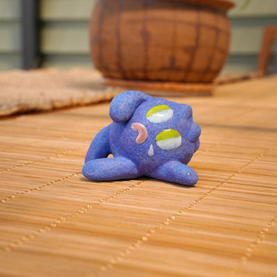 Small sculpture of a blueberry with a cute cartoon sad face - green eyes looking to the side and a frown with a single tear falling down. It lays on its side.