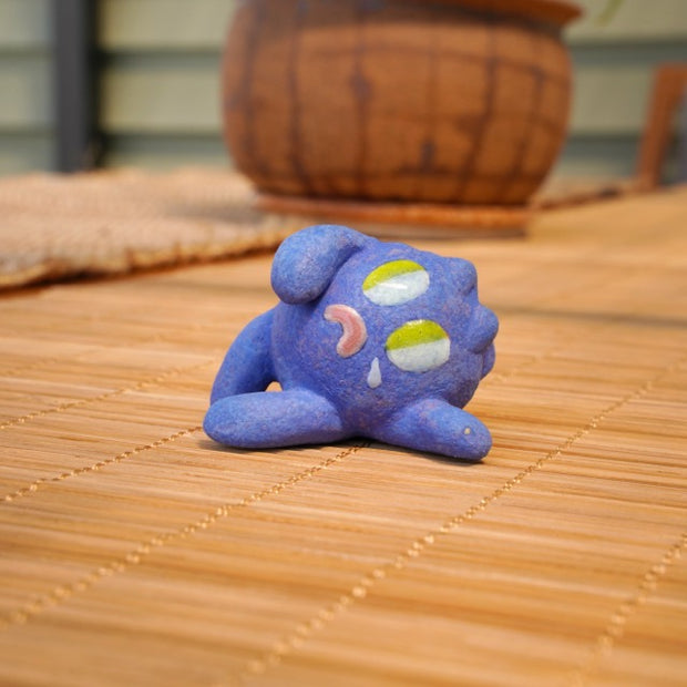 Small sculpture of a blueberry with a cute cartoon sad face - green eyes looking to the side and a frown with a single tear falling down. It lays on its side.