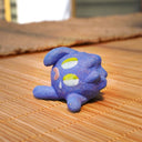 Small sculpture of a blueberry with a cute cartoon sad face - green eyes looking to the side and a frown with a single tear falling down. It lays on its side.