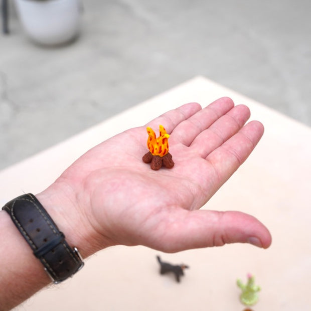 Hand holding a small model of a campfire with flames on a light-colored surface.