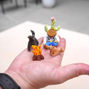Hand holding small figurines of a cowboy resting up against a cactus, a dog, and horse around a campfire.