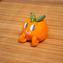 Small sculpture of an orange with 2 green leaves, and a cute cartoon face with purple eyes looking to the side and a simple smile.