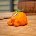 Small sculpture of an orange with 2 green leaves, and a cute cartoon face with purple eyes looking to the side and a simple smile. It lays on its "stomach" with its feet up and head resting in its hands.